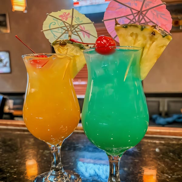Two vibrant cocktails, one orange and one blue-green, garnished with cherries, pineapple, and paper umbrellas.