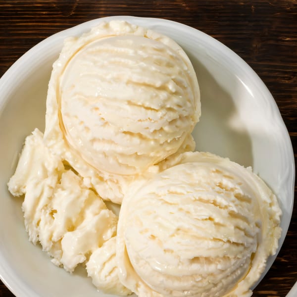 Three scoops of creamy vanilla ice cream in a white bowl on a dark wooden table.