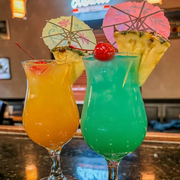 Two vibrant cocktails, one orange and one blue-green, garnished with cherries, pineapple, and paper umbrellas.